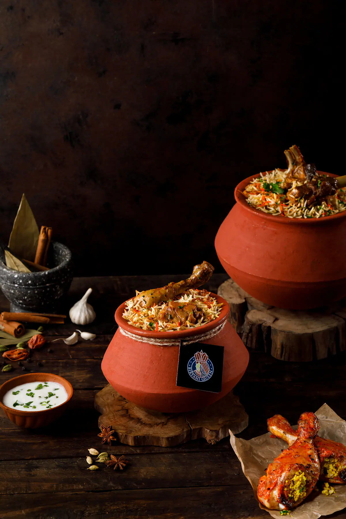 Two terracotta handis of dum biryani on wooden stands, with raita, whole spices and tandoori chicken